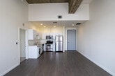 Located on the eighth floor of a building along Wilshire Boulevard in Los Angeles, Unit 814 showcases a distinctive industrial-loft aesthetic defined by its dramatically tall ceilings with exposed raw concrete beams. The open-plan living area is expansive and bathed in natural light through large arched windows dressed with sheer white curtains, offering views of the surrounding cityscape. Light-toned hardwood flooring runs throughout the main living space, enhancing the bright and airy atmosphere.

The kitchen area is integrated into the main living space and features white cabinetry and stainless steel appliances, including a full-size refrigerator. An in-unit washer is conveniently positioned nearby, adding practical functionality to the layout. The sleeping area is carpeted and separated from the main living space, continuing the soaring ceiling height with exposed concrete details overhead. Arched windows in the bedroom frame city views, and a mirrored sliding-door closet provides ample storage.

The bathroom is cleanly finished with a white vanity, a tub-shower combination, tile flooring, and a large mirror above the sink, maintaining a straightforward and functional design. Overall, the unit blends raw architectural character with clean, modern finishes throughout.