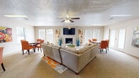 Located at 700 N Albert Pike Ave in Fort Smith, Arkansas, this single-floor space offers a well-organized, open layout encompassing a living room, dining area, and kitchen. The carpeted living room serves as the centerpiece, featuring a built-in fireplace flanked by colorful wall art, a wall-mounted television, and a ceiling fan with light fixture. Multiple windows along the perimeter flood the room with natural light, while a set of French doors provides access to the outdoors. A spacious sectional sofa anchors the room around a central coffee table atop a vibrant area rug, creating a welcoming gathering atmosphere. Adjacent to the living room, the dining area accommodates a round table with upholstered chairs and benefits from its own set of windows offering garden views. Colorful canvas artwork decorates the walls, adding warmth and personality to the space. The kitchen features white cabinetry, white appliances, tile flooring, and a peninsula with bar seating for two — ideal for casual dining or entertaining. A skylight and recessed lighting keep the kitchen bright and functional. Throughout the space, a textured ceiling, neutral walls, and consistent finishes contribute to a cohesive, comfortable ambiance.