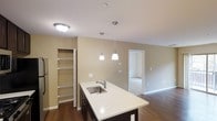 This single-floor space showcases a thoughtfully designed open-concept layout that integrates the kitchen and living areas into one cohesive environment. The kitchen serves as a central focal point, anchored by a large island with a crisp white countertop and an undermount stainless steel sink. Dark-toned cabinetry beneath the island provides a striking contrast against the light surfaces. Three pendant lights hang above the island, offering both functional task lighting and a modern design element that draws the eye upward.

Adjacent to the kitchen, a built-in pantry with adjustable wire shelving provides convenient and well-organized storage. The warm beige walls and rich hardwood flooring run throughout, creating a cohesive and inviting atmosphere. A flush-mount ceiling fixture adds additional ambient lighting to complement the pendant arrangement.

The open layout allows a clear sightline from the kitchen into the adjacent living space and through a doorway leading to a separate room beyond, suggesting flexible use of space throughout. The overall design balances practicality with clean, contemporary aesthetics, making the space both functional and visually appealing.