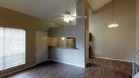 Located in Tyler, Texas, this two-story space offers a functional and open layout designed for comfortable everyday living. The main living area is anchored by vaulted ceilings that lend a sense of openness and height, complemented by a ceiling fan with a built-in light fixture that provides both airflow and ambient lighting. The flooring throughout the main level features wood-style planks in a warm, dark tone, adding a cohesive and modern aesthetic to the space.

A half-wall with a pass-through window connects the living area to the kitchen, creating an open sightline while maintaining a defined separation between the two spaces. The kitchen is visible through this opening, revealing white appliances, including a refrigerator, and cabinetry that provides ample storage. The pass-through counter also functions as a casual bar-style seating area, making it ideal for quick meals or conversation while cooking.

Neutral-toned walls in a warm taupe palette run throughout the main level, contributing to a calm and inviting ambiance. A white entry door is visible along one wall, indicating a convenient ground-floor entry point. With two floors total, the layout offers additional living or sleeping space above, providing a practical separation between common and private areas.