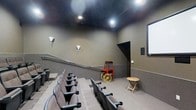 This single-floor home theater is designed to replicate the immersive atmosphere of a commercial cinema. The room features tiered, stadium-style seating arranged in rows of cushioned theater chairs, each equipped with armrests and cup holders, ensuring comfort during extended viewing sessions. The seating layout is elevated at the rear, providing unobstructed sightlines to the projection screen mounted on the front wall.

The space is finished with dark, moody tones throughout — a matte black ceiling absorbs light to minimize reflections, while the walls are painted in a deep taupe-gray, enhancing the cinematic ambiance. Recessed ceiling spotlights and wall-mounted sconces provide subtle, controllable illumination that complements the theater setting.

A standout decorative feature is a vintage-style red and yellow popcorn cart, adding a playful, classic movie-house charm to the room. A small folding side table is also present, offering a convenient surface for snacks or drinks. The rear of the room features a defined entryway with a dark-framed doorway, contributing to the space's cohesive, purpose-built design. Overall, this theater delivers a polished and functional private cinema experience.