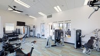 Located at 1420 Renaissance Drive in Longmont, Colorado, this single-floor fitness center offers a well-equipped and thoughtfully designed workout space. The room features a bright, open layout illuminated by recessed panel lighting and elevated ceilings with a clean white drop-tile finish, creating an airy and energizing atmosphere. Large windows line one wall, allowing natural light to flow in while providing views of the surrounding outdoor area. The fitness floor is outfitted with a comprehensive selection of exercise equipment, including cable and multi-function strength machines, seated resistance units, stationary cycling bikes, and cardio stations — catering to a wide range of workout preferences and fitness levels. A wall-mounted mirror enhances the sense of space and supports form-conscious training. The flooring features a vibrant multicolored striped carpet tile pattern in shades of teal, green, and yellow, adding a dynamic visual element to the space. Ventilation is well-integrated into the ceiling, ensuring comfort during intense workouts. A glass-paned door provides access to an adjacent area, contributing to a seamless and connected facility layout. Overall, the space is clean, modern, and functionally equipped to support a full fitness routine.