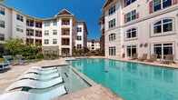 This outdoor amenity space is centered around a large, resort-style swimming pool with clear turquoise water and a gradual wading entry steps that ease into the shallow end — ideal for leisurely lounging. The pool deck is finished in natural travertine pavers that extend generously around the perimeter, providing ample space for sunbathing and outdoor relaxation. A separate circular spa is tucked to one side, offering a dedicated area for hydrotherapy. Numerous chaise lounge chairs are arranged throughout the deck, complemented by shaded seating areas with vibrant blue market umbrellas and covered pergola-style structures for those seeking relief from the sun. Lush landscaping with trees, shrubs, and potted plants adds greenery and a park-like ambiance to the setting. The courtyard is enclosed by multi-story buildings featuring a mix of white siding, stone veneer accents, arched windows, and red-accented balconies, creating a cohesive architectural backdrop. The space benefits from abundant natural light under open skies, making it a bright and inviting outdoor retreat suited for both active recreation and quiet relaxation.