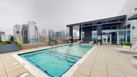 Located at 500 W Trade Street in the heart of uptown Charlotte, this rooftop amenity level offers an impressive collection of outdoor spaces designed for relaxation and socializing. The centerpiece is a resort-style swimming pool with multiple entry points, surrounded by rows of sleek lounge chairs and planters adding greenery against the modern building facade. A covered pergola area provides shaded outdoor seating alongside a dedicated grilling station, making it ideal for gatherings. Multiple distinct lounge zones are furnished with woven white-frame chairs and soft blue cushions, creating a coastal-inspired aesthetic set against warm wood-toned deck tiles. A covered clubroom with floor-to-ceiling glass walls opens directly onto the deck, offering interior seating that connects seamlessly to the outdoors. The rooftop also features open dining areas with white bistro tables and chairs positioned to take full advantage of the panoramic views. From virtually every vantage point on the deck, residents enjoy sweeping, unobstructed views of the Charlotte skyline, with the city's towers rising dramatically as a backdrop. The overall design blends contemporary architecture with approachable, resort-inspired comfort, creating a versatile outdoor destination above the city.