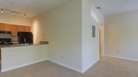 This single-story space presents a straightforward, functional layout with a clean, neutral aesthetic. The interior is finished with warm beige walls and light-colored carpet flooring throughout, creating a cohesive and understated look. A textured ceiling adds subtle visual interest overhead, while white baseboards provide a crisp contrast to the soft wall tones.

A defining feature of the main living area is the pass-through counter that connects to the kitchen, offering a practical and open link between the two spaces. The kitchen area, partially visible from the main room, includes dark appliances and a countertop ledge. This pass-through design promotes an open flow between the living and kitchen areas.

A central wall partition helps define the layout, with a recessed electrical panel mounted on its surface — a practical and accessible utility feature. An HVAC ceiling vent ensures climate control throughout the space. A hallway beyond the partition suggests additional rooms or storage areas.

Overall, the space offers a clean, move-in-ready environment with a simple and practical design, well-suited for comfortable everyday living.