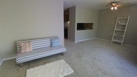 This single-floor space presents an open-plan layout that seamlessly connects the living area to the kitchen. The living room is generously sized, finished with soft wall-to-wall carpeting in a neutral beige tone that contributes to a warm and understated ambiance. A chevron-patterned futon sofa anchors the seating area, accompanied by a plush white area rug that adds texture to the floor space.

A partial dividing wall with a pass-through window separates the living area from the kitchen, creating a sense of definition between the two zones while maintaining an open feel. The kitchen is compact and functional, featuring dark cabinetry, a stovetop, and under-cabinet lighting that provides focused illumination to the workspace.

The walls throughout are finished in a consistent light grey-beige tone, lending the space a cohesive and calm aesthetic. Baseboards are clean and white, adding a crisp contrast to the carpeted floors. The overall design is simple and functional, with a neutral palette that offers flexibility for a variety of interior styling preferences.