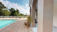 This single-story outdoor space showcases a well-maintained community pool area designed for relaxation and recreation in a warm Florida-style setting. The centerpiece is a generously sized rectangular swimming pool with clear turquoise water, accessible via built-in entry steps. Surrounding the pool is a broad, textured paver deck finished in earthy tones of tan and rust, offering ample space for lounge chairs and outdoor seating arrangements positioned around the pool's perimeter.

The space is framed by a white metal safety fence that encloses the entire pool area, providing both security and an open, airy feel with views of the surrounding mature trees, palms, and lush green landscaping beyond the boundary. Tropical ornamental plants, including striking dracaena, add vibrant greenery along the deck's edge.

Adjacent to the pool deck is a covered patio area supported by substantial stucco-finished columns with arched architectural detailing, characteristic of Mediterranean-influenced design. This shaded corridor features a smooth concrete floor and white-framed windows with louvered blinds, offering a cool retreat from the sun. The overall ambiance is bright, open, and inviting, well-suited to outdoor leisure under the expansive Florida sky.