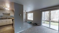 This single-floor residence offers a thoughtfully laid-out living space with a clean, neutral aesthetic throughout. The main living area is anchored by plush carpeting that transitions to smooth laminate hardwood-style flooring, clearly delineating different functional zones within the open floor plan. Warm taupe-toned walls are complemented by crisp white trim and baseboards, creating a cohesive and inviting atmosphere.

Natural light plays a central role in the space, with a large picture window fitted with horizontal blinds and a full-width sliding glass door that opens to an outdoor area, offering views of lush greenery beyond. The sliding door allows for abundant daylight to fill the room throughout the day. A ceiling fan with a light fixture overhead provides both ventilation and additional illumination.

A partial wall partition subtly divides the living area from the adjacent kitchen, which is visible to the side and features traditional cabinetry and countertops. The textured ceiling adds a classic architectural touch. A wall-mounted heating unit ensures year-round comfort. Overall, the space presents a functional and well-lit single-level layout suited for comfortable everyday living.