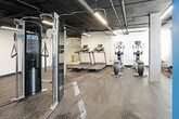 This fitness center offers a comprehensive workout environment designed to accommodate a wide range of exercise routines. The space features an industrial aesthetic, with exposed ceiling ductwork and structural concrete columns that lend a raw, urban character throughout. Durable rubber flooring covers the entire area, providing both safety and functionality underfoot.

The cardio zone is equipped with multiple treadmills and elliptical machines, positioned near a window that allows natural light to filter in, creating a welcoming atmosphere for sustained workouts. A mural of a city skyline adorns the nearby wall, adding a distinctive visual element to the space.

The strength training area houses a well-stocked dumbbell rack with a broad range of weights, supported by a cable machine for versatile resistance training. A dedicated functional training section features a bench press, a medicine ball rack stocked with assorted balls in various sizes, stability balls, and a full-length wall mirror that enhances spatial depth and supports form monitoring.

Bright linear LED lighting runs throughout the ceiling, ensuring clear visibility across all workout zones. The color scheme contrasts bold teal accent walls in the functional training area against clean white walls elsewhere, creating distinct zones within the open floor plan. Sanitation stations are conveniently placed to maintain cleanliness throughout the facility.