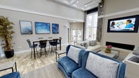 This thoughtfully designed single-floor communal space offers an inviting and sophisticated atmosphere centered around a cohesive blue and neutral color palette. The lounge is anchored by soaring tray ceilings with recessed lighting that enhance the sense of openness and grandeur throughout the room. Large floor-to-ceiling windows along one wall flood the interior with abundant natural light, creating a bright and welcoming environment at any time of day.

The space is divided into distinct functional areas that cater to a variety of activities. A casual seating area features a plush blue velvet sofa and a cream-toned couch, arranged around a central coffee table and accented with decorative throw pillows. Adjacent to this is a bar-height dining area furnished with sleek dark bistro tables and matching chairs, ideal for informal gatherings or individual use. Blue abstract artwork adorns the walls, reinforcing the room's cohesive aesthetic.

The flooring features a combination of tile and area rugs that define each zone while maintaining a unified visual flow. Glass double doors provide a seamless connection to an outdoor area, further enhancing the airy feel of the space. Decorative stone or tile wall paneling adds a refined, textural element near the windows, contributing to the overall polished and modern character of this well-appointed lounge.