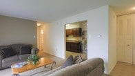 This single-floor residence offers a straightforward, functional layout with a comfortable living room serving as the central gathering space. The living area features wall-to-wall carpeting in a neutral tone, providing a warm foundation for the room. A plush sofa and armchair are arranged around a light wood coffee table accented with a small potted plant, creating an inviting atmosphere. The walls are painted in soft, neutral shades, contributing to a clean and understated aesthetic.

An open passageway connects the living room directly to the kitchen, allowing natural sightlines between the two spaces. The kitchen is outfitted with dark espresso-toned cabinetry that extends to the upper and lower portions of the walls, offering generous storage. A double sink sits beneath the cabinetry, and the countertops provide ample workspace. A decorative metal shelving unit adds a functional accent near the kitchen entrance.

A hallway visible from the living area leads further into the residence, suggesting additional rooms beyond the main living space. Overhead lighting in both the living area and kitchen ensures consistent illumination throughout. The overall ambiance is calm, practical, and well-organized.