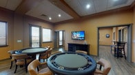 This well-appointed game room offers a diverse range of recreational amenities within a single, open layout. The space is divided into distinct activity zones, each designed to encourage social interaction and leisure. Warm amber-toned walls and wood-look flooring establish a welcoming, pub-inspired atmosphere throughout.

The billiards area anchors the main floor, featuring a full-sized pool table illuminated by an industrial-style pendant light fixture. A dartboard mounted on the wall and framed artwork add character to the surrounding space. Nearby, a shuffleboard table provides additional gaming options.

The card room area accommodates multiple round poker tables with padded leather edges and built-in cup holders, surrounded by comfortable upholstered chairs. A wall-mounted flat-screen television and a dark wood media console serve as a focal point in this zone.

A casual dining lounge features banquette seating along one wall, paired with dark wood café-style tables and chairs. Pendant lighting overhead and a series of framed vintage automobile photographs lend the area a distinctive, stylish character.

Large windows fitted with horizontal blinds allow natural light to filter throughout, offering views of the surrounding exterior. Recessed and track lighting ensure the space remains well-lit during evening use.