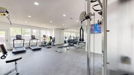 This fitness center is organized across two connected rooms, offering a comprehensive workout environment designed to support a wide range of training styles. The main room is bright and open, benefiting from large windows that fill the space with natural light and provide views of the surrounding area. It features durable rubber flooring throughout and recessed ceiling lighting. Cardio equipment, including treadmills and an elliptical machine, lines the windowed wall, while a full-length mirror along one side enhances the sense of space. A wall-mounted flat-screen TV keeps members engaged during workouts. A rack of dumbbells and a weight bench provide strength training options, and a multi-function cable machine anchors the room for versatile resistance training. The second room serves as a dedicated TRX Training Zone, housing an extensive wall-mounted rack stocked with kettlebells, medicine balls, and suspension training systems. A plyo box and additional accessories make this area ideal for functional and high-intensity training. Stability balls, medicine balls, and yoga mats are also available for stretching and core work. Together, the two rooms deliver a thorough, well-rounded fitness facility with a clean, modern aesthetic.