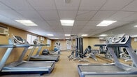 This fitness center provides a spacious, single-floor exercise facility designed to accommodate a wide range of workout routines. The room features a practical drop-panel ceiling with recessed fluorescent lighting that ensures consistent, even illumination throughout the space. Neutral-toned walls and a durable carpet flooring contribute to a clean, functional aesthetic.

The cardio section is well-stocked, featuring multiple treadmills, elliptical trainers, stair climbers, and both upright and recumbent stationary bikes from notable brands including Matrix and Precor. Complementing the cardio equipment is a multi-station cable strength training machine, along with a weight bench and free weights, offering users a comprehensive full-body workout experience.

Full-length mirrors line portions of the walls, visually expanding the space and allowing users to monitor their form during exercise. Natural light filters in through windows along one side of the room, while a dedicated access door provides a clearly marked entry and exit point. Safety features include a mounted fire extinguisher and visible exit signage.

The layout is thoughtfully organized, with cardio machines arranged in rows and strength equipment positioned to maximize the use of available floor space, ensuring comfortable circulation throughout the room. Posted fitness center rules and a security camera reflect an environment maintained with user safety and order in mind.