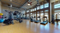 The Avenue 900 fitness center is a well-appointed, single-floor facility designed to support a full range of workout routines. The space is immediately distinguished by its soaring ceilings, which are fitted with an industrial-style exposed grid system, ceiling fans, and track lighting that ensures consistent illumination throughout. A generous bank of floor-to-ceiling windows lines one wall, flooding the interior with natural light and offering open views of the surrounding outdoors, creating an energizing atmosphere for exercise. The flooring throughout is a warm-toned hardwood, providing a clean and durable surface suited for both equipment use and open floor activities. The cardio section is well-stocked with multiple treadmills, elliptical trainers, and stationary bikes, all positioned to take advantage of the natural light from the windows. The strength-training area features a robust selection of Hoist cable machines, selectorized weight stations, benches, and free weights, including kettlebells and medicine balls. A heavy punching bag and large wall-mounted mirrors further enhance the training options. Two flat-screen televisions are mounted on the walls for entertainment. The facility is accessed through double glass doors marked with the Avenue 900 branding, offering a welcoming and professional entry point.