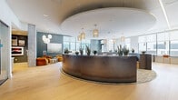 Located at 6945 E Chenango Ave in Denver, CO, this single-floor commercial space offers a well-appointed and visually cohesive environment across six distinct areas. The centerpiece of the space is a dramatic, curved reception desk clad in dark brushed metal, elegantly set beneath a coffered circular ceiling detail. Woven rattan pendant lights hang overhead, complementing the warm-toned light hardwood flooring that flows throughout.

The lounge and collaborative seating areas are defined by a lush living moss wall interspersed with vertical wood slat panels, introducing strong biophilic design elements that create a calming, nature-inspired atmosphere. Curved banquette seating in deep navy pairs with light wood accent chairs and round pedestal tables, all anchored by a soft teal-toned area rug. Floor-to-ceiling windows along the perimeter flood the space with natural light and frame dynamic views of the surrounding Denver cityscape.

Additional areas include semi-private enclosed rooms, likely suited for consultations or focused work, visible along one wing of the floor plan. Touches of greenery, including snake plants and succulents positioned along the reception counter, reinforce the space's commitment to a fresh, organic aesthetic. The overall ambiance is professional yet inviting, blending contemporary design with warm, natural materials.