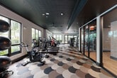 Located at 105 NE Sixth Ave in Delray Beach, Florida, this contemporary fitness center offers a well-appointed, single-level space designed to accommodate a comprehensive range of workout activities. The facility's interior is defined by a striking dark textured ceiling punctuated by recessed lighting, which creates a sleek, high-end ambiance throughout. A bold scalloped-pattern carpet in tones of charcoal, taupe, and cream covers the floor, serving as a distinctive design element that ties the space together visually.

Large windows line multiple walls, allowing abundant natural light to filter in while providing views of lush tropical landscaping — a nod to the South Florida setting. The cardio zone features rows of treadmills, ellipticals, stair climbers, and stationary bikes, each equipped with personal display screens. A dedicated strength training area houses multi-station cable machines, selectorized equipment, and an extensive free weight section with a full dumbbell rack and benches. A large flat-screen display is prominently mounted for entertainment or instructional use.

A functional training zone includes a power rack, punching bag, medicine balls, stability balls, and kettlebells, complemented by glass-partitioned areas for added spatial flexibility. The overall design balances modern aesthetics with practical fitness functionality.