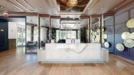 Located at 4310 Dunlavy St in Houston's Montrose neighborhood, this sophisticated two-floor space serves as a striking common area and lobby. The ground floor welcomes visitors through floor-to-ceiling glass entry doors, revealing polished travertine tile flooring that extends throughout. A sleek marble-fronted reception desk anchors the main lobby, set against a dramatic dark accent wall adorned with decorative circular art pieces. An ornate framed ceiling mural adds a refined touch overhead, while large windows invite abundant natural light and views of the surrounding greenery. A comfortable seating area with plush sofas and accent chairs provides an inviting lounge space. The lobby connects seamlessly to two hallways, one of which features a bold large-scale stained glass artwork reproduction, brass pendant lighting, and warm wood-toned tile flooring. A mirrored ceiling hallway leads to a stainless steel elevator and a grand staircase accented with brass railings and a striking sculptural chandelier. The upper floor expands the space considerably, housing additional hallways with wainscoting, panel molding, a beverage station with dark cabinetry, and a resident mailbox area. Throughout, the design balances classic architectural detailing with contemporary art and fixtures, creating a cohesive and upscale atmosphere.