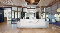 Located at 4310 Dunlavy St in Houston's Montrose neighborhood, this sophisticated two-floor space serves as a striking common area and lobby. The ground floor welcomes visitors through floor-to-ceiling glass entry doors, revealing polished travertine tile flooring that extends throughout. A sleek marble-fronted reception desk anchors the main lobby, set against a dramatic dark accent wall adorned with decorative circular art pieces. An ornate framed ceiling mural adds a refined touch overhead, while large windows invite abundant natural light and views of the surrounding greenery. A comfortable seating area with plush sofas and accent chairs provides an inviting lounge space. The lobby connects seamlessly to two hallways, one of which features a bold large-scale stained glass artwork reproduction, brass pendant lighting, and warm wood-toned tile flooring. A mirrored ceiling hallway leads to a stainless steel elevator and a grand staircase accented with brass railings and a striking sculptural chandelier. The upper floor expands the space considerably, housing additional hallways with wainscoting, panel molding, a beverage station with dark cabinetry, and a resident mailbox area. Throughout, the design balances classic architectural detailing with contemporary art and fixtures, creating a cohesive and upscale atmosphere.