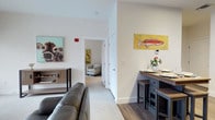 This one-bedroom unit is thoughtfully arranged across a single floor, maximizing its efficient use of space. The open layout connects the living and dining areas seamlessly, creating a cohesive flow throughout the home. The living area features light-toned carpet and accommodates comfortable seating, while colorful wall art adds personality to the neutral, white-painted walls. A hallway leads directly to the bedroom, which is visible from the main living space and benefits from the unit's open sightlines.

The dining area is defined by a dark-toned built-in table with matching stools, offering a compact yet functional spot for meals. The contrast between the dark hardwood-style flooring in the dining zone and the carpeted living and bedroom areas helps delineate each space naturally. White trim and clean-lined doorframes contribute to a crisp, modern aesthetic throughout.

The unit's light color palette, combined with its straightforward layout and practical built-in furnishings, creates a tidy and welcoming ambiance suited for everyday living.