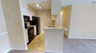 This single-floor residence offers a well-organized and functional layout designed to maximize everyday convenience. The kitchen is the heart of the space, outfitted with black appliances including a gas range with oven, built-in microwave, and refrigerator. White cabinetry provides ample storage both above and below the countertops, while a secondary cabinet nook adds additional space for pantry items or display. A peninsula breakfast bar with a rounded edge extends from the kitchen, creating a natural divide between the kitchen and the adjacent living area, and offering casual seating or additional prep space. The stainless steel sink is neatly integrated into the countertop, positioned to overlook the living space. The kitchen features durable tile flooring, while the connecting living area transitions to soft carpeting, adding warmth and comfort underfoot. Recessed track lighting illuminates the kitchen, ensuring a bright and practical cooking environment. The overall color palette — warm beige walls, white trim, and neutral flooring — creates a clean, cohesive ambiance throughout the space. A doorway visible beyond the kitchen hints at additional rooms, contributing to the overall sense of a connected and livable floor plan.