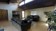 This single-story home makes an immediate impression with its dramatic vaulted ceiling, where rich, dark-stained wood paneling and exposed beams draw the eye upward and lend a warm, rustic character to the interior. A ceiling fan is centrally positioned within the living area, complementing the clerestory windows that allow natural light to pour in from above, brightening the space throughout the day.

The open living area is generously proportioned, with light-toned hardwood flooring that contrasts beautifully against the darker ceiling tones. The layout comfortably accommodates ample seating, creating a welcoming environment for both relaxing and entertaining. A sliding glass door provides direct access to the outdoors, while additional windows along the walls ensure a bright and airy atmosphere.

The living space flows naturally into a kitchen and dining area, where bar-height seating at a counter creates an informal dining option and keeps the social connection between rooms intact. The open sightlines between the living and kitchen areas reinforce the home's cohesive, open-concept design.

Overall, the home balances rustic architectural details with practical, comfortable living, making it an inviting and functional single-floor residence.
