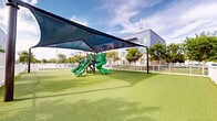 Located at 15350 Southwest 136th Street in Miami, Florida, this outdoor recreational space is designed to provide a safe, engaging environment for children and community members. The centerpiece of the area is a multi-feature playground structure equipped with multiple slides, climbing elements, and activity panels, all finished in vibrant green tones. Overhead, a large tensile shade sail canopy — supported by sturdy dark metal posts — stretches across the play zone, providing meaningful sun protection in Miami's warm climate.

The ground throughout the space is covered in lush artificial turf, offering a clean, low-maintenance, and cushioned surface ideal for active play and open recreation. The entire area is enclosed by a classic white picket fence, clearly defining the boundaries of the space and contributing to a safe, welcoming atmosphere.

Mature trees are planted along the perimeter, providing additional natural shade and greenery that softens the surroundings. The backdrop features a modern multi-story residential building with contemporary architectural detailing, including stone accents, large windows, and private balconies. A single-story community or amenity building is also visible, contributing to the cohesive design of the overall development. The space is bright, open, and thoughtfully designed for outdoor enjoyment.