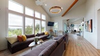 Located at 6347 SW Canyon Ct in Portland, Oregon, this single-floor community space showcases a contemporary, open-concept design characterized by soaring vaulted ceilings with exposed wooden beams and warm wood-look flooring throughout. The layout flows seamlessly from zone to zone, creating a welcoming and versatile environment well-suited for gathering and socializing.

The kitchen area is a standout feature, equipped with stainless steel appliances, a large center island topped with a bright white quartz countertop, a full white subway tile backsplash, and a prominent stainless steel range hood suspended from above. Pendant lighting and bar-height seating along the island add both function and style.

The lounge area is generously proportioned, anchored by large sectional sofas and defined by floor-to-ceiling windows that flood the space with natural light and frame sweeping views of the surrounding Pacific Northwest treeline. A circular gold-toned pendant light serves as a dramatic focal point beneath the vaulted ceiling.

Additional seating nooks with accent chairs and small tables provide more intimate settings throughout. Glazed sliding doors connect to outdoor areas, further enhancing the bright, airy atmosphere. The overall aesthetic is modern and clean, balancing warm natural materials with sleek, contemporary finishes.