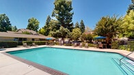 Located at 2593 Millcreek Dr in Sacramento, CA, this outdoor recreational area is a well-appointed amenity space centered around a generously sized swimming pool with clear, blue-green water. The pool deck is finished in a neutral concrete surround, providing ample room for lounge chairs and seating arrangements that invite relaxation under the California sun. Blue market umbrellas are positioned throughout the deck, offering shaded spots for residents and guests. A separate hot tub spa is situated nearby, fitted with stainless steel handrails and surrounded by its own seating area, including high-top tables and chairs ideal for casual gatherings. The entire outdoor space is enclosed by a secure metal fence, ensuring privacy and safety. Mature trees, including towering redwoods and a stately palm, frame the perimeter of the area, creating a lush, park-like atmosphere and natural shade. Manicured shrubs and landscaping add to the inviting aesthetic. In the background, residential buildings with gabled rooflines and wood siding are visible, complementing the natural surroundings. The overall setting is bright, open, and well-maintained, offering a resort-like outdoor experience.
