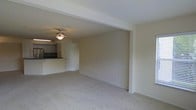 This single-floor space features an open-concept layout that connects the living area to the kitchen, creating a functional and cohesive floor plan. The living area is finished with plush, light-toned wall-to-wall carpeting that adds warmth underfoot, while the neutral cream-colored walls provide a clean, versatile backdrop throughout. A ceiling fan with an integrated light fixture is centrally positioned overhead, offering both air circulation and ambient lighting to the space. Structural elements include a prominent overhead beam that defines the transition between the living area and the rest of the unit, adding subtle architectural character. The kitchen is accessible from the main living space and features a breakfast bar counter, providing a casual dining or workspace option. Dark countertops offer a visual contrast to the lighter surroundings. A white door near the kitchen leads to an additional area, likely a utility or storage room. Electrical outlets are conveniently placed along the baseboards. The overall ambiance is straightforward and move-in ready, with a neutral palette that accommodates a wide range of furnishing styles.