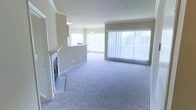 This single-floor residential space offers a welcoming and functional layout designed for comfortable everyday living. The main living area is generously proportioned, finished with soft wall-to-wall carpeting that adds warmth underfoot. Crown molding along the ceiling provides a refined architectural detail that elevates the overall aesthetic.

A standout feature of the living room is the fireplace, framed in white with a tiled hearth, serving as a natural focal point for the space. Adjacent to the living area, a pass-through counter connects to what appears to be a kitchen or dining area, allowing for an open and communicative floor plan ideal for entertaining or daily interaction.

Natural light is a defining characteristic of this space, with large sliding glass doors or floor-to-ceiling windows dressed in vertical blinds lining one full wall of the living area. These expansive windows not only brighten the interior with abundant daylight but also offer views to the outdoors, creating a seamless visual connection to the exterior. A pendant light fixture overhead adds a decorative lighting element to the dining or kitchen transition zone.

The neutral color palette throughout — soft beige walls, light carpet, and white trim — creates a clean, versatile backdrop that accommodates a wide range of interior styles and furnishings.