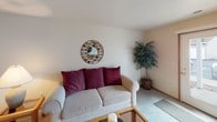 This single-floor guest suite offers a well-organized layout designed for comfortable short- or long-term stays. The living area features neutral-toned carpet, a textured ceiling, and soft ambient lighting, creating a relaxed atmosphere. A decorative round mirror serves as a focal point on the main wall, complementing the understated, warm aesthetic throughout the suite.

Adjacent to the living area, the dining space accommodates a four-seat table beneath a ceiling fan with an integrated light fixture, providing both comfort and functionality. The entry door and a secondary interior door are conveniently accessible from this area.

The kitchen is efficiently arranged in an L-shaped configuration, featuring white cabinetry with warm wood trim, light countertops, a stainless steel sink, and white appliances including a full-size refrigerator and dishwasher. Tile flooring adds practicality to this workspace.

The carpeted bedroom offers a quiet retreat, benefiting from natural light through a window with blinds and ample space for furnishings including a dresser and bedside storage. A hallway connects the bedroom to the full bathroom, which includes a tub/shower combination with a curtain, a single vanity, and a closet door for added storage.