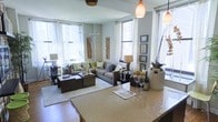 This single-floor space showcases an open-concept layout that seamlessly connects the kitchen and living areas, creating a cohesive and functional environment. The kitchen features a generous granite countertop with warm speckled tones, providing ample prep and entertaining space. Pendant lights hang above the counter, adding a refined touch to the area.

The living room is well-appointed with large windows on multiple walls, flooding the space with natural light and offering views of the surrounding urban landscape. The generous ceiling height enhances the sense of openness and airiness throughout. A comfortable sectional sofa in neutral gray tones anchors the seating area, complemented by coordinating accent pillows in green and white. Dark wood coffee and side tables add contrast and sophistication.

Decorative elements include framed wall art, sculptural branches, and carefully arranged accessories that contribute to a polished, contemporary aesthetic. The overall ambiance is modern and inviting, balancing clean architectural lines with warm, layered décor. The abundant natural light, urban views, and open flow between spaces make this a highly livable and visually appealing environment.