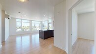 This single-floor residence showcases a clean, contemporary design throughout, with light hardwood flooring and crisp white walls creating a bright, cohesive aesthetic. The open-concept living and kitchen area serves as the heart of the space, where floor-to-ceiling windows and transom windows along multiple walls flood the interior with abundant natural light and frame expansive outdoor views of the surrounding neighborhood and skyline. The kitchen is outfitted with dark cabinetry, stainless steel appliances including a range, microwave, and refrigerator, and a large center island with a white countertop and integrated sink, offering generous workspace and casual seating potential.

The bedrooms continue the light hardwood flooring and feature large picture windows that draw in natural light and outdoor views. Each bedroom includes closet space, with at least one room offering a walk-in closet with built-in shelving. A wall-mounted mini-split system provides individual climate control. The bathrooms are finished with neutral tile work, including a glass-enclosed walk-in shower with subway tile surround and a separate tub with a handheld showerhead. Recessed lighting throughout reinforces the unit's modern, polished character.