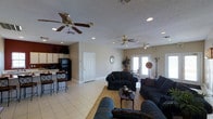 This single-floor property offers a generous open-concept layout that seamlessly integrates the living, dining, and kitchen areas into one cohesive space. The living area is well-appointed with multiple plush sofas arranged around a central coffee table, creating a comfortable and inviting gathering space. Light-toned ceramic tile flooring extends throughout, contributing to a bright and airy atmosphere. Two ceiling fans are strategically positioned across the ceiling to ensure consistent airflow throughout the space. Recessed lighting complements the natural light that filters in through a set of French doors at the far end of the room, which also provide access to the exterior. A dining area with a bistro-style table and chairs sits adjacent to the living space, offering a casual dining option. The kitchen, partially open to the main living area, features light wood cabinetry, a black refrigerator, and a breakfast bar with counter-height seating — ideal for quick meals or entertaining. The textured ceiling and neutral warm-toned walls throughout provide a clean, understated backdrop that ties the entire space together cohesively.