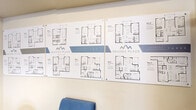 Located at 30 Nevada Place in St. Albert, Alberta, this single-floor office space serves as the on-site management and leasing hub for the Nevada Place property. The space is thoughtfully organized into two functional zones within one open room. The reception and client-facing area greets visitors with comfortable blue upholstered seating arranged around a glass coffee table, alongside a prominent wall display of Nevada Place floor plans — an informative reference for prospective residents. The Nevada Place logo is boldly featured on the wall, reinforcing the professional branding of the space. The workstation area is equipped with a large L-shaped desk featuring integrated overhead cabinetry, dual monitors, and ample workspace for administrative tasks. A rolling ergonomic chair, a multi-drawer filing cabinet, and additional wall-mounted storage cabinets provide excellent organizational capacity. A compact beverage refrigerator and small appliances are neatly integrated along one wall, adding convenience for staff. The space is finished with neutral beige walls, textured carpet tile flooring, and a textured ceiling. Lighting is provided by a combination of overhead fluorescent fixtures and a ceiling fan, while a wall-mounted split air conditioning unit ensures year-round comfort. A window allows for natural light to brighten the workstation area.