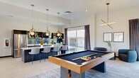 This expansive single-floor amenity space offers a sophisticated blend of leisure and social gathering areas, combining a living room and game room across a generously sized, open layout. Light-toned hardwood-style flooring flows throughout, tying the spaces together with a clean, contemporary aesthetic.

The game room serves as a central hub of activity, anchored by a full-size pool table with a rich wood-and-slate finish, complemented by a shuffleboard table and a collection of classic arcade cabinets. A geometric-patterned accent wall adds visual depth, while large windows dressed in floor-to-ceiling drapes flood the area with natural light and offer views of the surrounding greenery. Sculptural pendant lighting overhead adds a modern, artistic touch.

Adjoining the game room is a well-appointed bar-style lounge area featuring a long white countertop island with bar seating, dark cabinetry with open shelving, a tiled backsplash, and stylish pendant fixtures. This space transitions seamlessly into the living room, which features a large sectional sofa, accent chairs, and curated wall art.

A standout feature is a glass-panel folding door wall that opens to an outdoor patio with mountain views, inviting abundant sunlight and a strong indoor-outdoor connection. The overall ambiance is modern, vibrant, and welcoming.