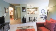 This single-floor residence offers a practical and inviting open-concept layout that connects the living and kitchen areas seamlessly. The living space is finished with wall-to-wall carpeting and features a colorful area rug that anchors the seating area, where a warm red armchair adds a pop of color. A decorative black accent cabinet with a table lamp provides a charming touch along one wall, accompanied by a framed mirror above.

The kitchen is defined by a raised pass-through bar counter, lined with dark bar stools that invite casual dining and conversation. Upper cabinets and open shelving above the counter display decorative items and greenery, with pendant lighting and a flush-mount fixture illuminating the space. A cheerful "Be Happy" sign adds a personal touch to the kitchen shelf display.

A hallway visible beyond the living area leads to additional rooms, with a doorway hinting at further living quarters. The walls throughout are painted in a soft sage green, contributing to a calm and cohesive atmosphere. The textured ceiling and neutral tones create a comfortable, functional environment well-suited for everyday living.