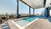 Located in the heart of Salt Lake City at 201 E 300 S, this rooftop amenity deck offers a sophisticated urban retreat with sweeping, unobstructed views of both the Wasatch Mountain range and the downtown skyline. The generously sized space is defined by its contemporary architectural design, featuring bold cylindrical columns, floor-to-ceiling glass curtain walls, and large-format porcelain tile flooring throughout.

At the center of the outdoor deck sits a long lap pool with crystal-clear water, flanked by rows of lounge chairs ideal for relaxation. Adjacent to the pool, a spacious hot tub provides a more intimate setting, finished with elegant tiled surrounds. The seamless integration of indoor and outdoor areas is achieved through expansive glass walls that allow natural light to flood interior zones while maintaining visual continuity with the outdoor pool deck.

An interior lounge area adjacent to the pool features warm, upholstered seating positioned directly against the floor-to-ceiling glass, framing dramatic cityscape views. Scattered seating clusters with side tables and lounge chairs are thoughtfully arranged across the terrace, offering multiple vantage points to take in the mountain and city panoramas. The overall ambiance is modern, open, and resort-inspired, making this rooftop deck a standout amenity in downtown Salt Lake City.