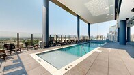 Located in the heart of Salt Lake City at 201 E 300 S, this rooftop amenity deck offers a sophisticated urban retreat with sweeping, unobstructed views of both the Wasatch Mountain range and the downtown skyline. The generously sized space is defined by its contemporary architectural design, featuring bold cylindrical columns, floor-to-ceiling glass curtain walls, and large-format porcelain tile flooring throughout.

At the center of the outdoor deck sits a long lap pool with crystal-clear water, flanked by rows of lounge chairs ideal for relaxation. Adjacent to the pool, a spacious hot tub provides a more intimate setting, finished with elegant tiled surrounds. The seamless integration of indoor and outdoor areas is achieved through expansive glass walls that allow natural light to flood interior zones while maintaining visual continuity with the outdoor pool deck.

An interior lounge area adjacent to the pool features warm, upholstered seating positioned directly against the floor-to-ceiling glass, framing dramatic cityscape views. Scattered seating clusters with side tables and lounge chairs are thoughtfully arranged across the terrace, offering multiple vantage points to take in the mountain and city panoramas. The overall ambiance is modern, open, and resort-inspired, making this rooftop deck a standout amenity in downtown Salt Lake City.
