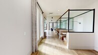 This thoughtfully designed single-floor community space offers a generous and versatile layout that seamlessly blends social and functional areas. Light oak hardwood flooring flows throughout, unifying the various zones under soaring double-height ceilings that amplify the sense of openness.

The main lounge area is anchored by bold blue accent chairs arranged around a central rug, with a large wall-mounted display and floor-to-ceiling windows that flood the room with natural light and offer views of the surrounding community. Dramatic pendant lighting descends from the high ceilings, adding a refined, modern touch.

A communal dining and co-working area features a long bar-height table with industrial-style seating, framed by expansive windows draped in soft linen curtains. Semi-private booth seating, enclosed within sleek black steel and glass frames, lines the hallway corridor — ideal for focused work or quiet conversation.

A glass-partitioned private meeting room with a round table and modern chairs provides an enclosed workspace with outdoor views. Near the entry, built-in bookshelves, mailbox banks, and a marble-accented service counter add practical functionality with elevated design detail. The hallway connects all areas fluidly, creating an intuitive, welcoming flow throughout the entire space.