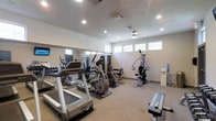 This fitness center offers a comprehensive workout environment designed to accommodate a range of exercise needs. The space is laid out on a single floor, providing a clean and accessible layout for users. Recessed lighting throughout the ceiling, combined with a row of clerestory windows along the upper walls, ensures the room is well-lit, supplemented by ample natural light.

The cardio section features multiple treadmills and an elliptical trainer, positioned on protective floor mats. A multi-station cable and pulley strength machine occupies one side of the room, offering versatile resistance training options. A flat-screen TV is mounted on the wall for entertainment during workouts. A dedicated dumbbell rack with a selection of free weights lines one wall, supported by a large wall-to-wall mirror that visually expands the space and allows users to monitor their form.

The flooring consists of a neutral carpet with rubber matting beneath equipment for added stability. A water cooler is conveniently positioned near the strength equipment. The overall ambiance is functional and tidy, with a neutral color palette of grey and white that maintains a clean, focused atmosphere suited to fitness activity.