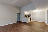 This single-floor residence offers a well-designed layout that balances open communal spaces with private living quarters. The living area is immediately impressive, featuring soaring vaulted ceilings with a textured finish and rich, dark wood-style flooring that runs throughout the main areas. Natural light filters in through windows at the entry, creating a bright and welcoming atmosphere. The space flows seamlessly from the living area toward the kitchen, which is accessible via a half-wall peninsula that doubles as additional counter space. The galley-style kitchen is well-appointed with dark gray shaker cabinetry, granite-style countertops, black appliances including a refrigerator, range, and dishwasher, and track lighting overhead. A dedicated laundry or utility closet is accessible directly from the kitchen corridor. The bedrooms are carpeted for comfort and feature neutral gray walls, ample space, and walk-in closets with wire shelving. The bathroom includes a vanity with dark cabinetry, a granite-style countertop, a large mirror, and a combined tub and shower. Throughout the residence, consistent neutral tones, clean white trim, and practical finishes create a cohesive and functional living environment.
