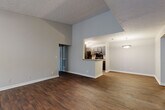 This single-floor residence offers a well-designed layout that balances open communal spaces with private living quarters. The living area is immediately impressive, featuring soaring vaulted ceilings with a textured finish and rich, dark wood-style flooring that runs throughout the main areas. Natural light filters in through windows at the entry, creating a bright and welcoming atmosphere. The space flows seamlessly from the living area toward the kitchen, which is accessible via a half-wall peninsula that doubles as additional counter space. The galley-style kitchen is well-appointed with dark gray shaker cabinetry, granite-style countertops, black appliances including a refrigerator, range, and dishwasher, and track lighting overhead. A dedicated laundry or utility closet is accessible directly from the kitchen corridor. The bedrooms are carpeted for comfort and feature neutral gray walls, ample space, and walk-in closets with wire shelving. The bathroom includes a vanity with dark cabinetry, a granite-style countertop, a large mirror, and a combined tub and shower. Throughout the residence, consistent neutral tones, clean white trim, and practical finishes create a cohesive and functional living environment.