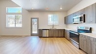 This three-story home in Bluffdale, Utah offers a well-organized layout spread across three distinct levels. The main living floor features a modern kitchen finished with wood-tone cabinetry, white quartz countertops, light hardwood-style flooring, and a full suite of stainless steel appliances including a refrigerator, range, microwave, and dishwasher. Natural light flows in through multiple windows, and a rear door provides convenient access to the exterior. An open living area adjoins the kitchen, highlighted by light hardwood flooring and a staircase with clean white railings. The upper floor hosts the bedrooms, each carpeted and bathed in natural light through large windows with blinds. The spacious primary bedroom features dual corner windows, while additional bedrooms offer ample room and bright outlooks. The four bathrooms are finished with wood-tone vanities, white countertops, light hardwood-style flooring, and full tub-shower combinations. The lower level includes an unfinished basement with insulated walls and concrete floors, offering significant potential for future development, along with a utility room for mechanical systems. Throughout the home, a neutral color palette and consistent modern finishes create a clean, cohesive aesthetic.