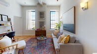 This thoughtfully designed single-floor residence offers a harmonious blend of classic architectural details and modern finishes throughout. Warm hardwood floors flow seamlessly across the living area, kitchen, and bedroom, creating a cohesive and inviting atmosphere.

The living room is a welcoming focal point, anchored by a large statement artwork and flanked by a pair of wall sconces that cast a warm glow. Multiple windows with black-framed trim flood the space with natural light and offer pleasant outdoor views, while high ceilings lend an open, airy quality to the room.

The kitchen is efficiently laid out with sleek grey cabinetry, white countertops, and stainless steel appliances, including a range and dishwasher. A pendant light above the sink adds a touch of character, and the space is designed to maximize both function and style.

The bedroom is a calm retreat, featuring a mini-split climate control unit for year-round comfort, a closet with a sliding door, and curated artwork that enhances the serene ambiance.

The bathroom is finished with dark hexagonal mosaic tile flooring, crisp white cabinetry, and clean white panel doors. A framed artwork and neatly hung towels complete the polished look, with a shower-tub combination rounding out the practical amenities.