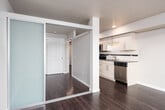This well-appointed single-floor unit offers a thoughtfully designed layout that balances functionality and modern style. Dark hardwood flooring runs throughout, creating a warm and cohesive aesthetic across every room.

The kitchen is a standout feature, showcasing crisp white cabinetry, dark countertops with a tile backsplash, and a full suite of stainless steel appliances including a range, dishwasher, microwave, and refrigerator. Recessed lighting and ample cabinet storage make this an efficient and attractive workspace that flows naturally into the open living area.

The living space is bright and open, anchored by a large floor-to-ceiling window that fills the room with natural light and frames views of the surrounding urban neighborhood. This generous window also connects the interior to the outdoor environment, offering a sense of elevation and openness.

The bedroom is a calm, neutral-toned retreat featuring the same rich hardwood flooring and a set of sliding panel closet doors with clean, paneled detailing — providing practical storage while maintaining a streamlined look.

The bathroom is finished with a wide vanity featuring dark countertops, a large wall mirror, light wood cabinetry below, and a shower/tub combination — offering a clean and functional design. Overall, the unit presents a modern, move-in ready space with consistent finishes throughout.