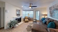 This single-floor space offers a thoughtfully designed living area that balances comfort and style. The living room serves as the heart of the home, anchored by a classic white-trimmed fireplace with a decorative mantle, adding warmth and character to the space. A large bay window configuration floods the room with natural light, framed by elegant curtains and horizontal blinds that allow for flexible light control. Light wood-toned flooring extends throughout, complementing the neutral, airy color palette of the walls and ceiling. A ceiling fan with integrated lighting provides both function and a finishing touch overhead, while recessed lighting adds to the bright, welcoming atmosphere. The room is generously proportioned, offering ample space for a full seating arrangement, including a plush sectional sofa accented with teal throw pillows, a cushioned armchair with ottoman, and layered area rugs that define the gathering space. A side door provides convenient exterior access, enhancing the functionality of the layout. Lush indoor greenery adds life and a natural element to the interior. Overall, the space presents a clean, inviting aesthetic well-suited for everyday living and entertaining.