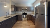 This single-floor residence is designed with a clean, modern aesthetic and a practical open-concept layout. The living area serves as the central hub of the space, featuring light-toned hardwood-style flooring, neutral gray walls, and a ceiling fan with integrated lighting. A peninsula counter connects the living area to the kitchen, enhancing the open flow between spaces.

The kitchen is well-equipped and functional, showcasing dark espresso-toned shaker cabinetry paired with granite-look countertops and a full suite of stainless steel appliances, including a refrigerator, range, built-in microwave, and dishwasher. Light wood-style flooring continues throughout, and ample upper and lower cabinetry provides generous storage.

The bedroom is generously sized and features plush carpeting, a ceiling fan, and sliding glass doors opening to a private balcony with outdoor views. A secondary room, also carpeted, offers additional flexibility. The bathroom maintains the same design language as the kitchen, with matching dark cabinetry, granite-look countertops, a large mirror, and hardwood-style flooring. Overall, the unit presents a cohesive and contemporary aesthetic throughout.