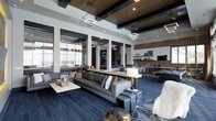 This stylish, single-floor lounge space showcases a refined contemporary design that blends comfort with upscale aesthetics. The open-plan layout is anchored by soaring ceilings finished with rich wood beam and plank detailing, contrasted against crisp white structural columns that define the space without enclosing it. Striking geometric pendant light fixtures suspend from above, casting a warm, modern glow throughout. Floor-to-ceiling windows line one wall, flooding the interior with natural light and offering expansive outdoor views that enhance the sense of openness. The main seating area features a generous sectional sofa in deep charcoal, complemented by a sleek marble-topped coffee table and coordinating upholstered ottomans — all resting atop rich navy-blue carpeting that grounds the room's cool, sophisticated palette. A central bar or reception counter with a light wood surface divides the lounge from a secondary area, which includes dining seating and a billiards table, pointing to a multi-functional entertainment focus. The back wall showcases a tiled feature wall with open shelving displaying curated décor, alongside a mounted flat-screen television. The overall ambiance is polished, modern, and inviting — ideal for socializing and relaxation.